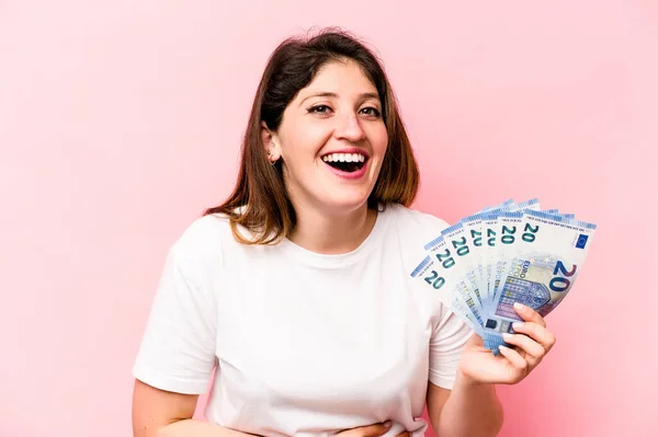 Young caucasian woman holding banknotes isolated on pink background laughing and having fun.