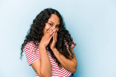 Mavi arka planda izole edilmiş genç İspanyol kadın bir şeye gülüyor, elleriyle ağzını kapatıyor..