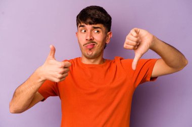 Mor arka planda izole edilmiş genç beyaz adam baş parmağını kaldırıyor ve baş parmağını aşağı gösteriyor, zor seçim kavramı.