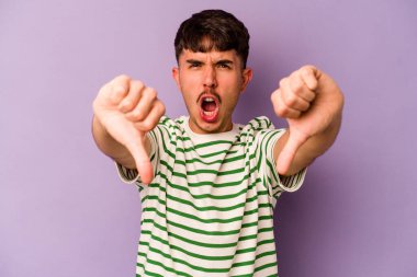Mor arka planda izole edilmiş genç beyaz adam baş parmağını aşağı gösteriyor ve hoşlanmadığını ifade ediyor..