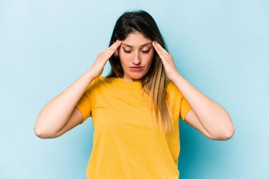 Mavi arka planda izole edilmiş genç beyaz kadın şakaklara dokunuyor ve baş ağrısı çekiyor..