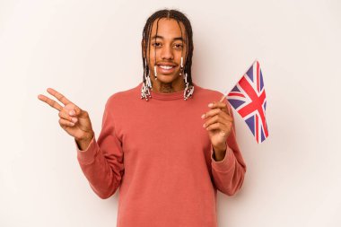 Beyaz arka planda izole edilmiş İngiliz bayrağı tutan genç Afrikalı Amerikalı adam neşeli ve kaygısız parmaklarıyla barış sembolü gösteriyor..