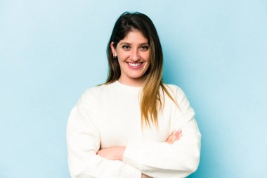 Mavi arka planda izole edilmiş genç beyaz kadın kendini güvende hissediyor, kararlılıkla kollarını kavuşturuyor..