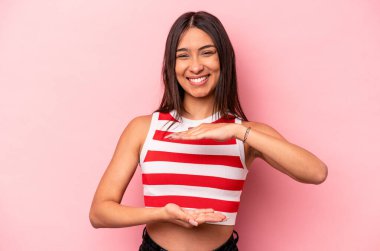 Pembe arka planda izole edilmiş genç İspanyol kadın iki eliyle bir şey tutuyor, ürün sunumu..