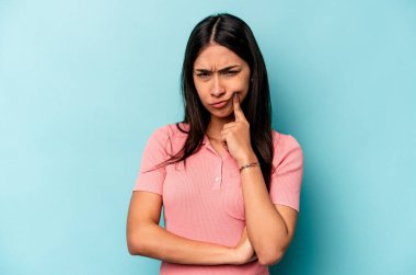 Genç İspanyol kadın mavi arka planda izole edilmiş. Mutsuz ve alaycı bir ifadeyle kameraya bakıyor..