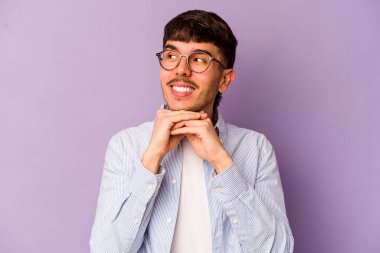 Mor arka planda izole edilmiş genç beyaz adam ellerini çenesinin altında tutuyor, mutlu bir şekilde yana bakıyor..
