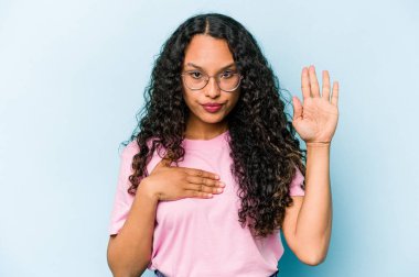 Genç İspanyol kadın mavi arka planda izole edilmiş yemin ediyor, elini göğsüne koyuyor..