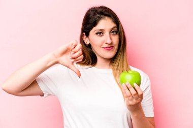 Elma tutan beyaz bir kadın pembe arka planda izole edilmiş, hoş olmayan bir jest gösteriyor, başparmakları aşağıda. Anlaşmazlık konsepti.