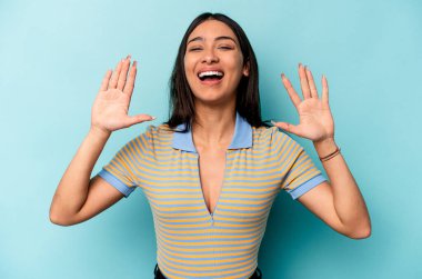 Genç İspanyol kadın mavi arka planda izole edilmiş. Hoş bir sürpriz bekliyor. Heyecanlı ve el kaldırıyor..