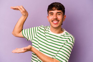 Mor arka planda izole edilmiş genç beyaz bir adam. Elinde işaret parmaklarıyla küçük bir şey tutuyor, gülümsüyor ve kendinden emin..