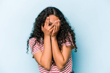 Mavi arka planda izole edilmiş genç İspanyol kadın kameraya parmaklarıyla göz kırpıyor, yüzünü kapatmaktan utanıyor..