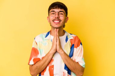 Sarı arka planda izole edilmiş genç beyaz bir adam el ele tutuşup ağız kenarında dua ediyor, kendinden emin hissediyor..