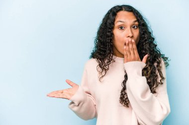 Mavi arka planda izole edilmiş genç İspanyol kadın avuç içi fotokopi alanını tutmaktan etkilenmiş..