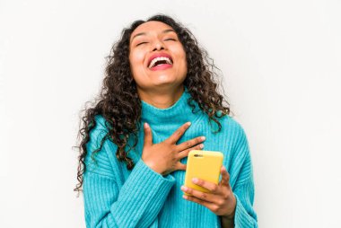 Beyaz arka planda cep telefonu tutan Latin kökenli genç bir kadın elini göğsünde tutarak kahkahalar atıyor..