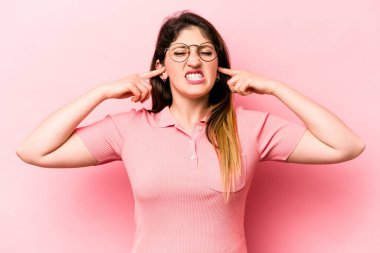 Pembe arka planda izole edilmiş genç beyaz bir kadın parmaklarıyla kulaklarını kapatıyor, gürültülü bir ortam yüzünden stresli ve çaresiz..