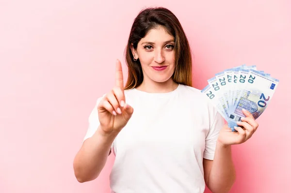 ピンクの背景に孤立した紙幣を持っている若い白人女性は指でナンバーワンを示す — ストック写真