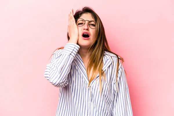 Pembe arka planda izole edilmiş genç beyaz kadın yorgun ve uykulu ellerini başının üstünde tutuyor..