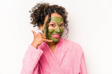 Beyaz arka planda izole edilmiş yüz maskesi takan genç İspanyol kadın parmaklarıyla bir cep telefonu çağrısı hareketi gösteriyor..