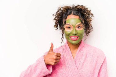 Beyaz arka planda soyutlanmış yüz maskesi takan genç İspanyol kadın gülümsüyor ve başparmağını kaldırıyor.