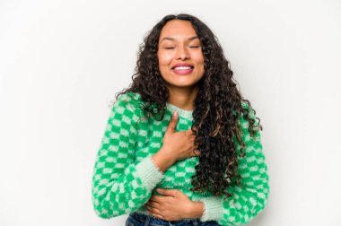 Beyaz arka planda izole edilmiş genç İspanyol kadın mutlu bir şekilde gülüyor ve ellerini karnının üstünde tutarak eğleniyor..