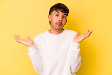 Sarı arka planda izole edilmiş genç beyaz adam sorgulama sırasında omuz silkiyor ve kuşkulanıyor..