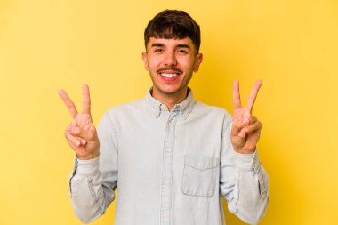 Sarı arka planda izole edilmiş genç beyaz adam zafer işareti gösteriyor ve gülümsüyor..