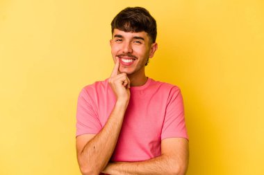 Sarı arka planda izole edilmiş genç, mutlu ve kendinden emin gülümseyen, elleriyle çenesine dokunan beyaz bir adam..