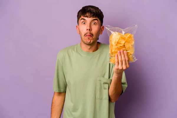 Jonge Spaanse Man Met Chips Geïsoleerd Paarse Achtergrond Haalt Schouders — Stockfoto