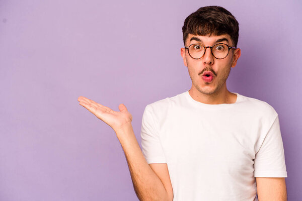 Young caucasian man isolated on purple background impressed holding copy space on palm.