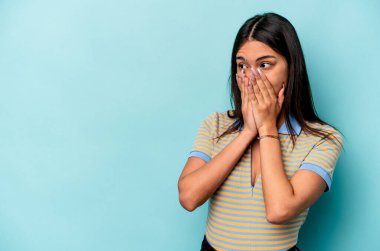 Genç İspanyol kadın mavi arka planda izole edilmiş. Düşünceli bir şekilde bir fotokopi odasına bakıyor. Ağzını eliyle kapatıyor..