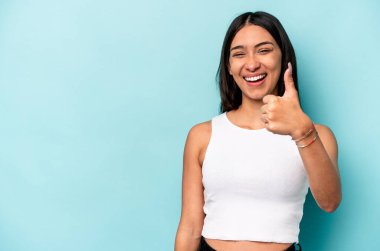 Mavi arka planda izole edilmiş genç İspanyol kadın gülümsüyor ve başparmağını kaldırıyor.