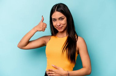 Mavi arka planda izole edilmiş genç İspanyol kadın karnına dokunuyor, nazikçe gülümsüyor, yemek yiyor ve tatmin oluyor..