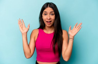 Genç İspanyol kadın mavi arka planda izole edilmiş zaferi ya da başarıyı kutluyor, şaşırmış ve şok olmuş..