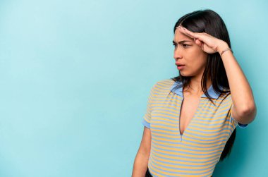 Mavi arka planda izole edilmiş genç İspanyol kadın uzaklara bakıyor ve elini alnına koyuyor..