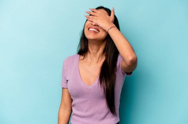 Mavi arka planda soyutlanmış genç İspanyol kadın elleriyle gözlerini kapatıyor, geniş bir gülümsemeyle bir sürpriz bekliyor..