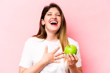 Pembe arka planda soyutlanmış bir elma tutan beyaz bir kadın yüksek sesle gülüyor ve elini göğsünde tutuyor..
