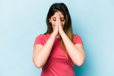 Mavi arka planda izole edilmiş genç beyaz bir kadın el ele tutuşup ağza yakın dua ediyor, kendinden emin..