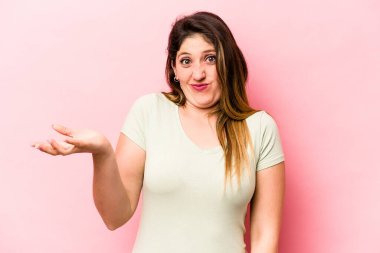 Pembe arka planda izole edilmiş genç beyaz kadın sorgulama sırasında omuz silkiyor ve şüphe duyuyor..