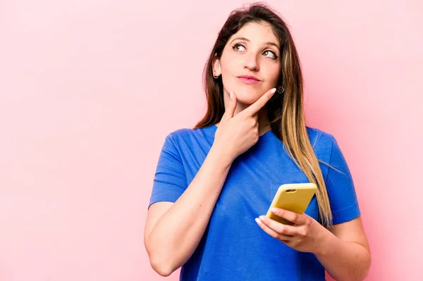 분홍색 배경에 고립된 휴대폰을 코카서스 여성은 의심스럽고 회의적 표현으로 바라본다 — 스톡 사진