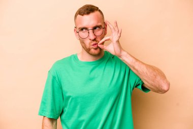 Beyazlı genç bir adam bej arka planda izole edilmiş ve parmakları dudaklarında bir sır saklıyor..