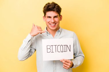 Sarı arka planda Bitcoin levhası tutan genç beyaz adam parmakları olan bir cep telefonu hareketi gösteriyor..