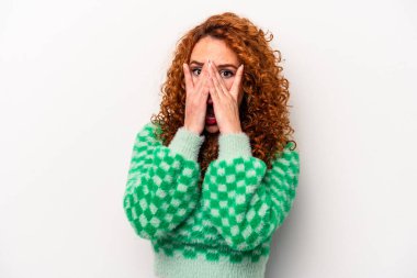 Beyaz arka planda izole edilmiş genç, beyaz bir kadın parmaklarının arasından korkmuş ve gergin bir şekilde göz kırpıyor..