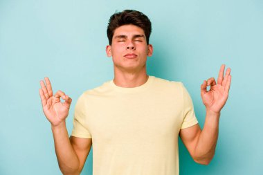 Mavi arka planda izole edilmiş genç beyaz adam yorucu bir günün ardından rahatlıyor. Yoga yapıyor..