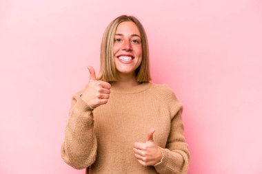 Pembe arka planda izole edilmiş genç beyaz kadın iki parmağını da kaldırıyor, gülümsüyor ve kendinden emin..