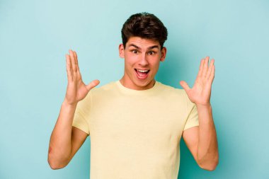 Mavi arka planda izole edilmiş genç beyaz bir adam zaferi ya da başarıyı kutluyor, şaşırıyor ve şok oluyor..