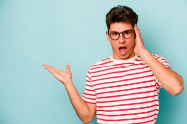 Mavi arka planda izole edilmiş genç beyaz adam, avuç içi fotokopi alanını elinde tutmaktan etkilenmiş..