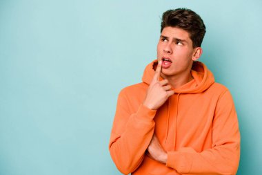 Mavi arka planda izole edilmiş genç beyaz adam şüpheli ve şüpheci bir ifadeyle yana bakıyor..