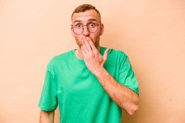 Beyaz tenli genç adam bej arka planda izole edilmiş şok olmuş, ağzı ellerle kaplanmış, yeni bir şey keşfetmeye hevesli..