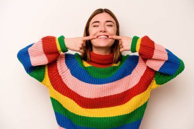 Beyaz arka planda izole edilmiş genç beyaz kadın gülümsüyor, parmaklarını ağıza doğrultuyor..