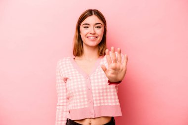 Pembe arka planda izole edilmiş genç beyaz kadın gülümseyerek beş numarayı parmaklarıyla gösteriyor..